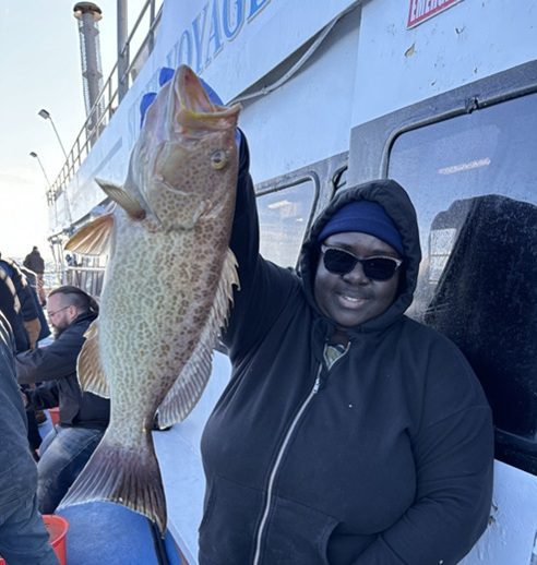 Person in sunglasses holding a large fish on a boat, smiling at the camera.